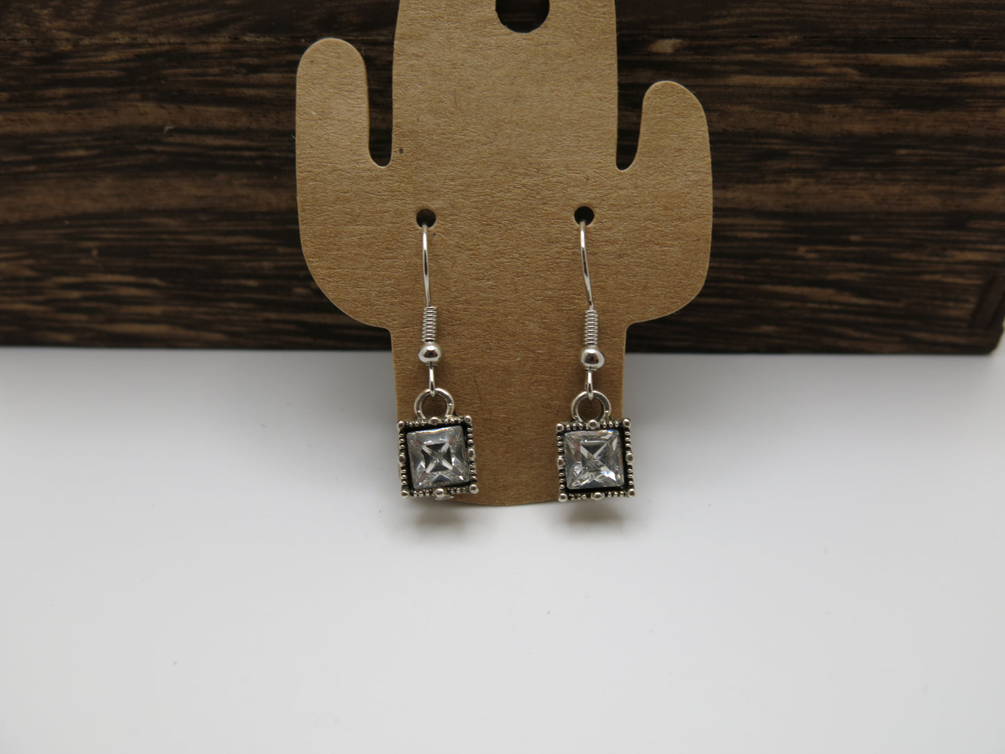 Silver square earrings on a cactus-shaped wooden display card against a wooden background.