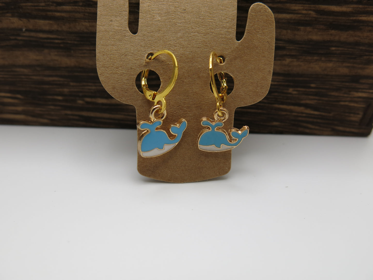 Gold earrings with blue whale designs on a cardboard backing against a wooden background