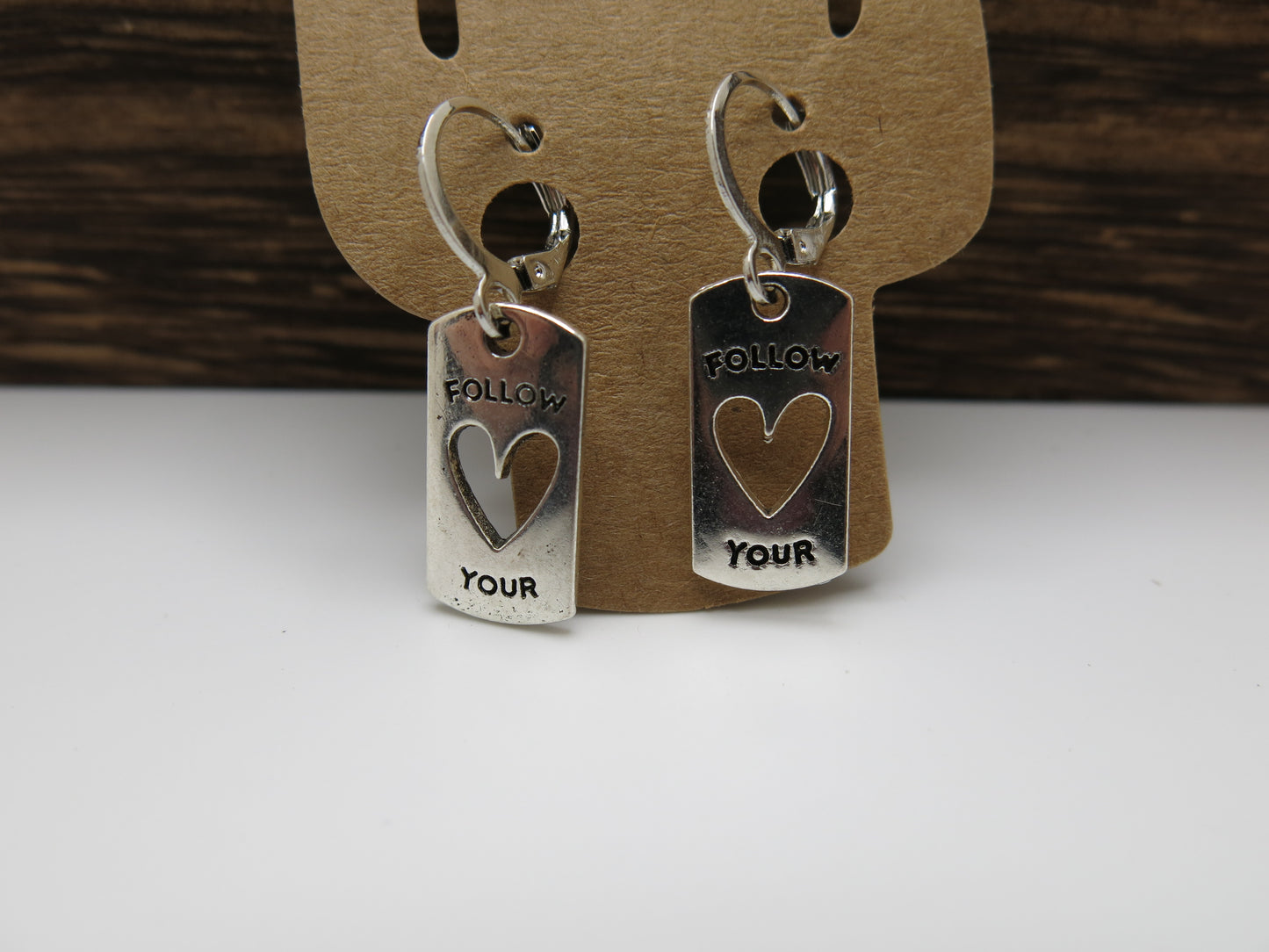 Silver tag earrings with 'Follow Your Heart' text on a brown card with wood background.