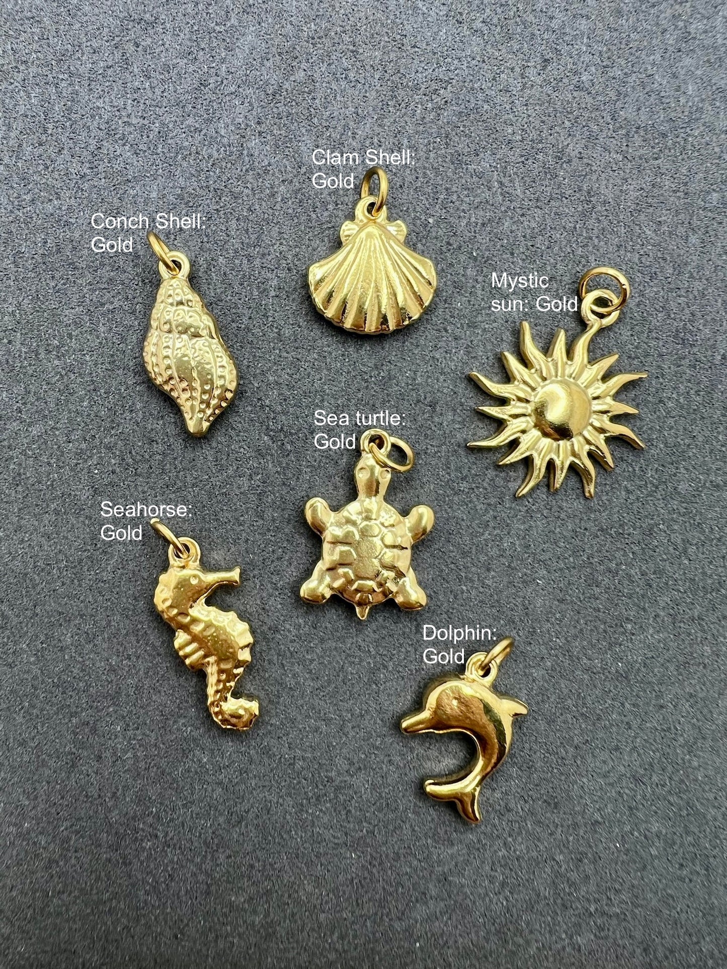 Set of gold charms including conch shell, clam shell, sea turtle, seahorse, dolphin, and mystic sun on a gray background.