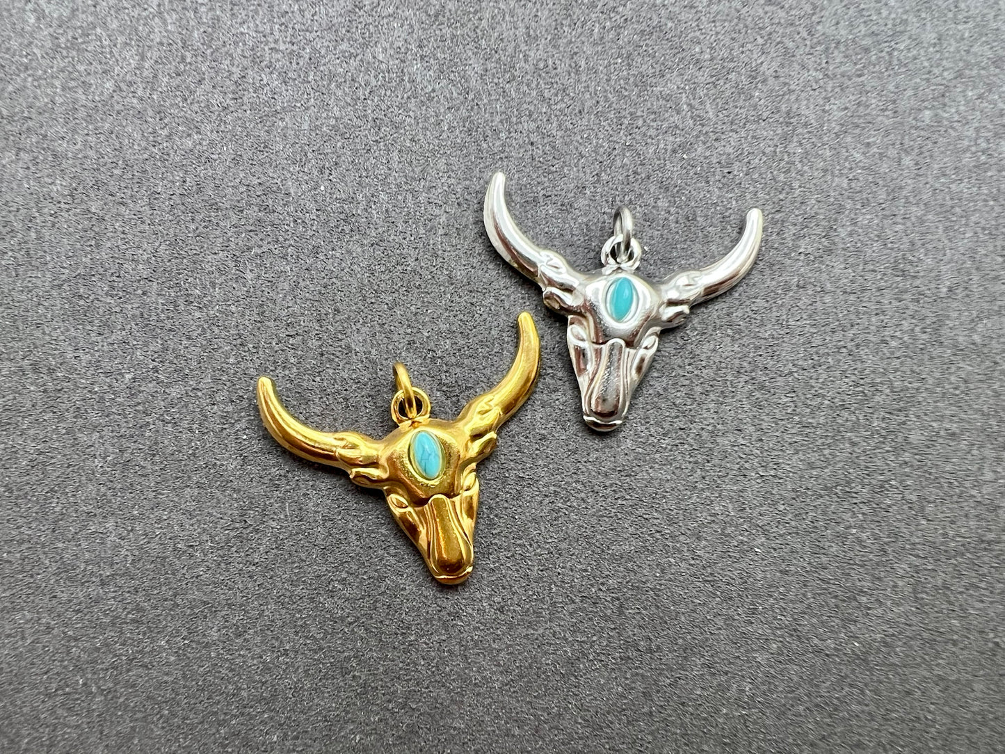 Two bull skull pendants, one gold and one silver, on a gray background.