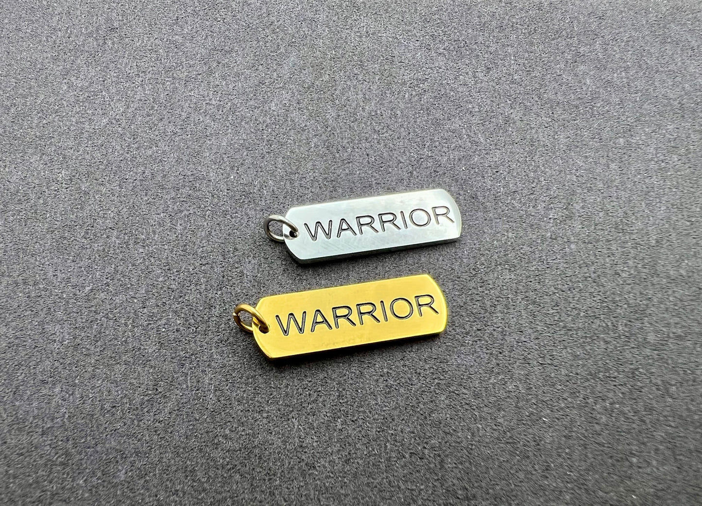 Two 'WARRIOR' tags, one gold and one silver, on a gray textured background.